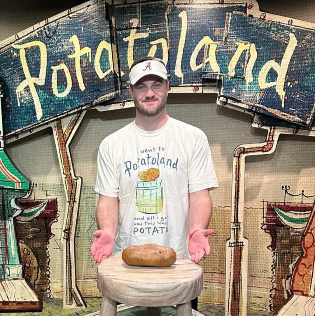 Potatoland Tee in Ivory worn by a man in a ball cap presenting a potato on a stool in front of the Potatoland sign