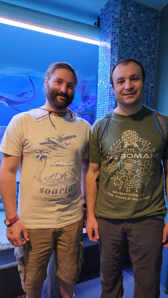 Two men posing in front of an aquarium, one wearing the Soarin' Around the World Tee in a light neutral color