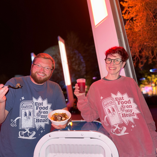 Disney Trash Can Sweatshirt in Denim and Crimson worn by a man and Kylie eating festival food at Epcot at night