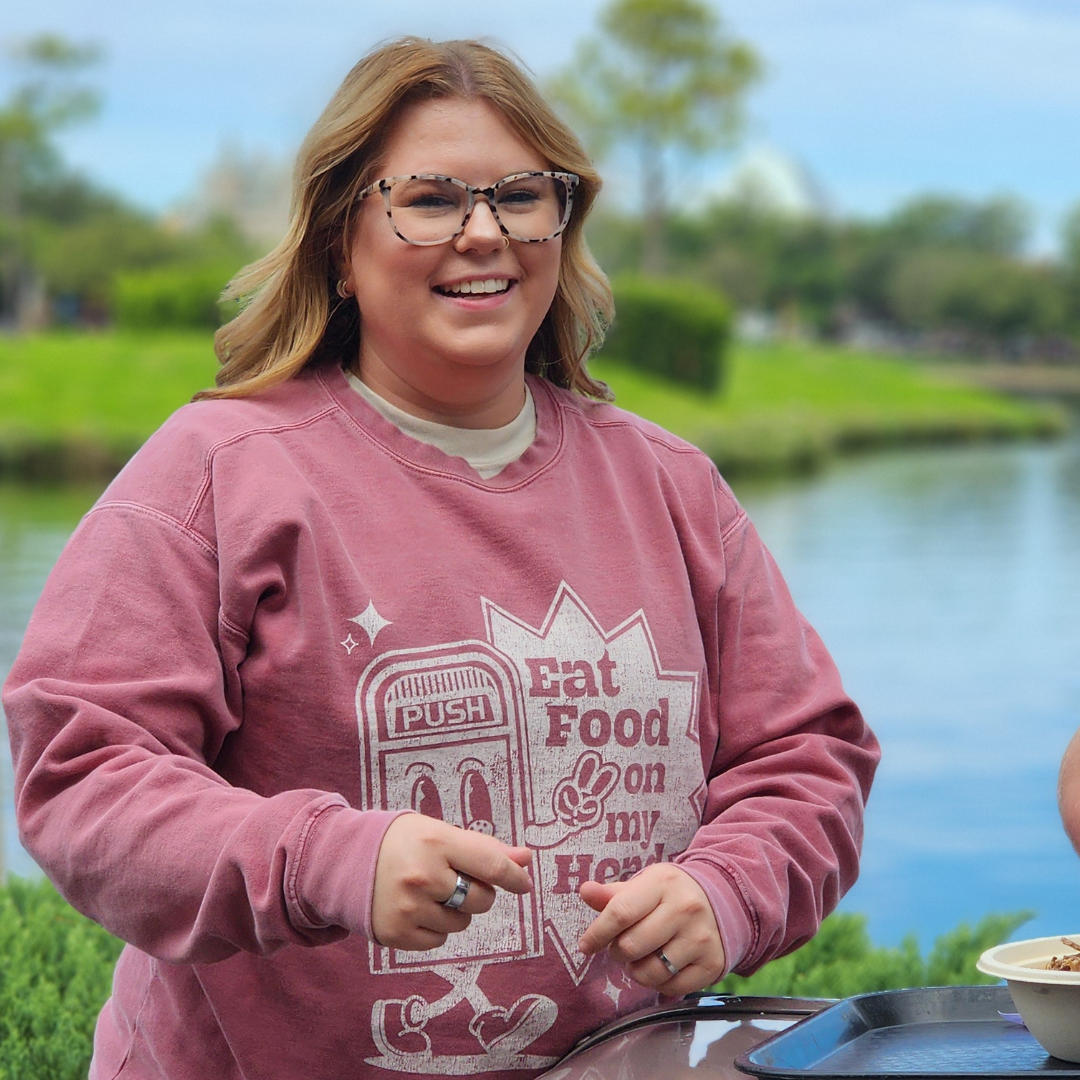 Disney Trash Can Sweatshirt in Crimson worn by a smiling woman beside the water at Epcot