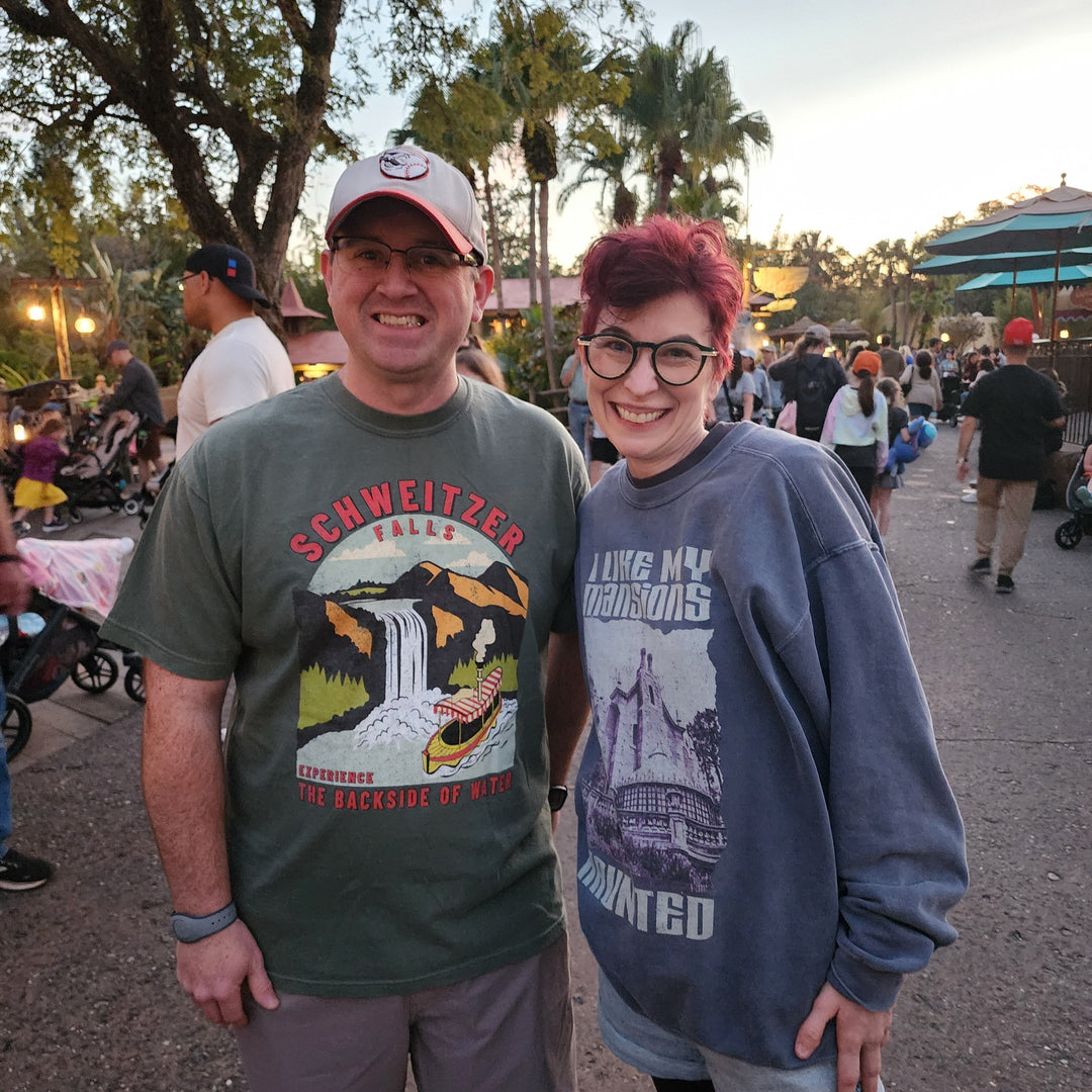 Man in the Schweitzer Falls Tee beside Kylie in her Haunted Mansion sweatshirt at Magic Kingdom at dusk