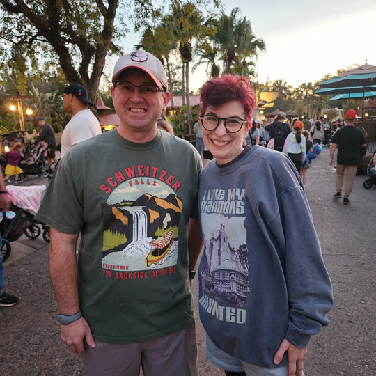 Man in the Schweitzer Falls Tee beside Kylie in her Haunted Mansion sweatshirt at Magic Kingdom at dusk
