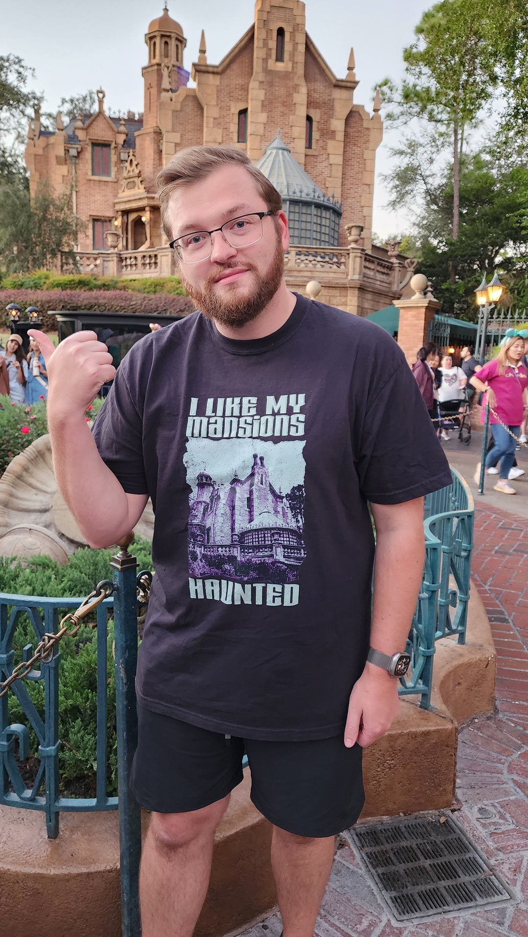 I Like My Mansions Haunted Tee in Black worn by a bearded man giving a thumbs up outside the Haunted Mansion at dusk