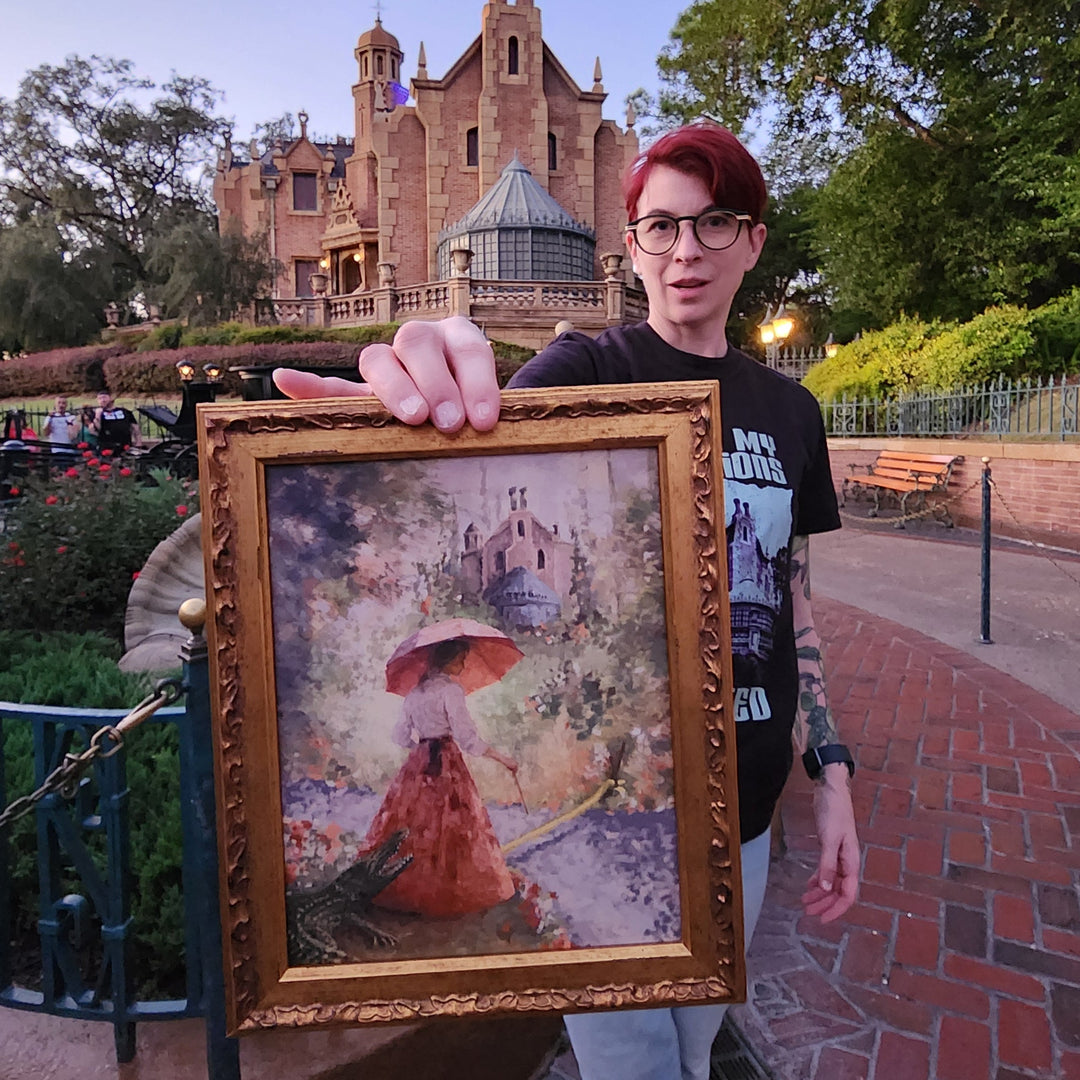 Artist Kylie Bodiya holding a framed picture with Sally Slater from the Haunted Mansion Stretching room with the haunted mansion in the background