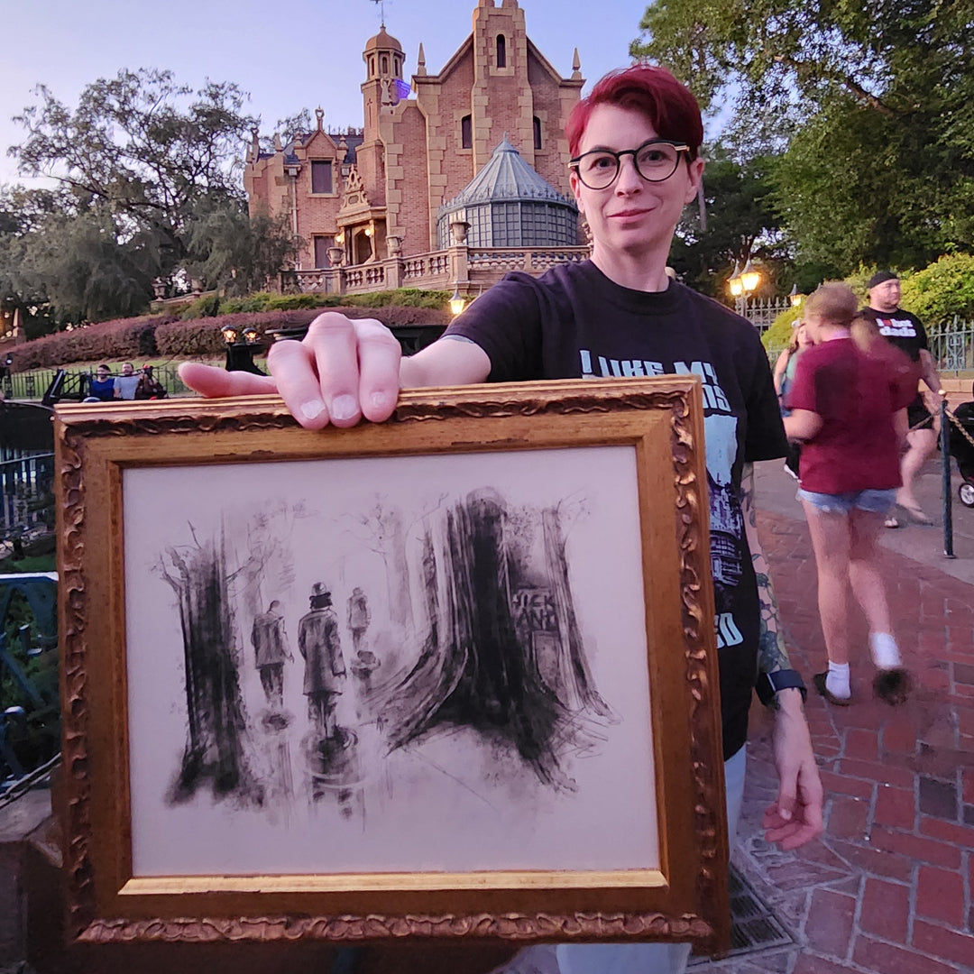 Artist Kylie Bodiya holding a framed picture with the Quicksand Trio from the Haunted Mansion Stretching room with the haunted mansion in the background
