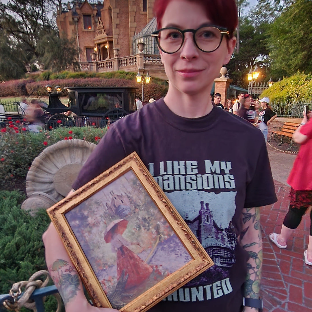 Kylie in glasses holding the gold-framed Sally Slater print at an angle outside the Haunted Mansion