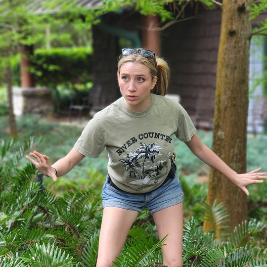River Country Tee in Bay worn by a woman crouching playfully among ferns and trees, like exploring ruins