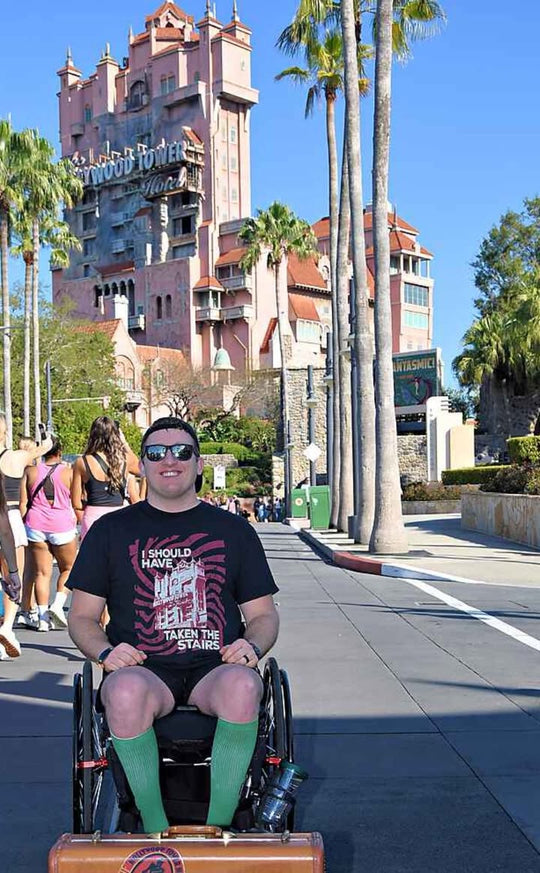 Man in a wheelchair wearing the Should Have Taken The Stairs Tee in Black in front of Tower of Terror on a sunny day