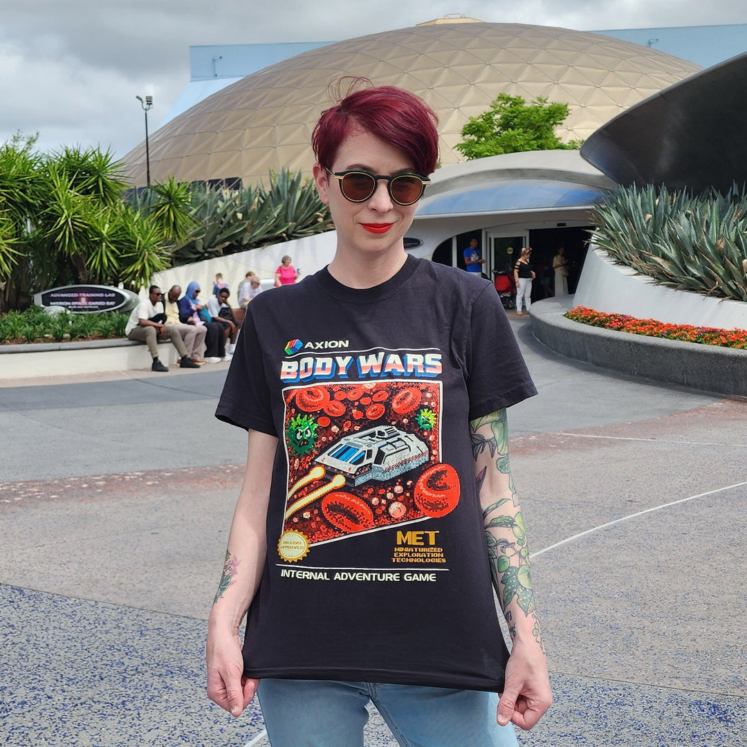 Body Wars Tee in Black worn by a woman with red hair and sunglasses in front of the former Wonders of Life pavilion