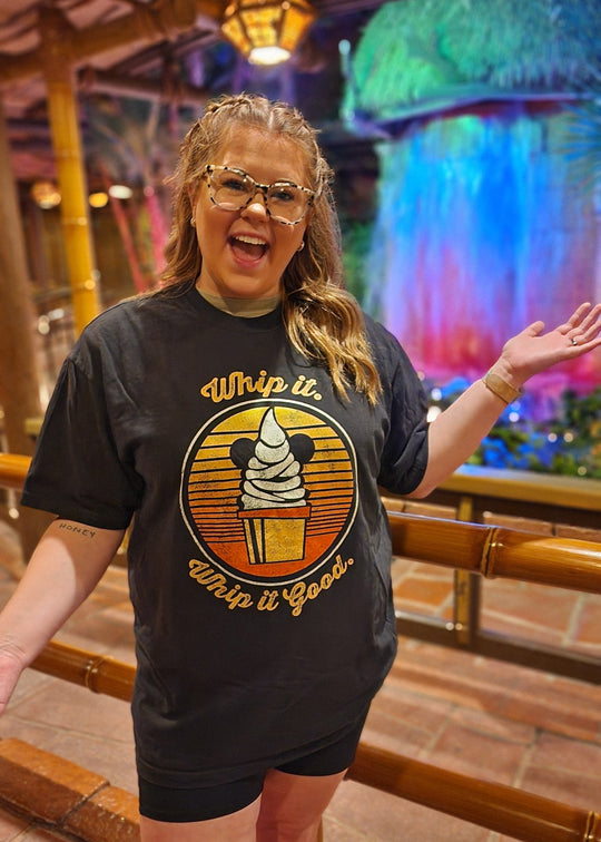Dole Whip Tee in Black worn by a smiling woman at a colorful tropical-themed restaurant with a lit waterfall