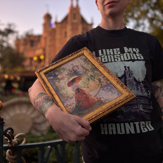 Framed Sally Slater print held against an I Like My Mansions tee outside the Haunted Mansion at dusk