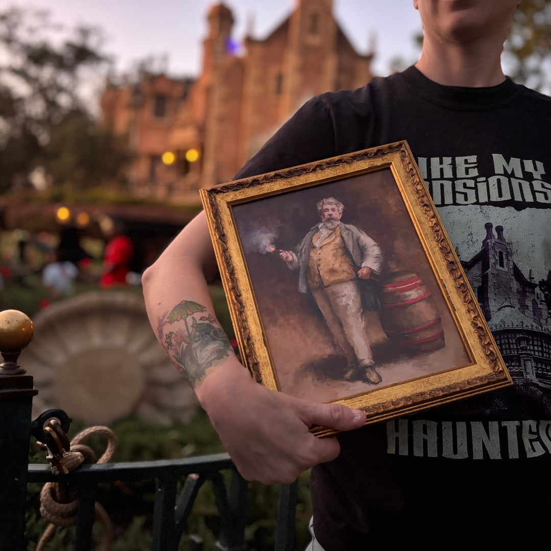 Framed Dynamo Fine Art Print held in front of a Haunted Mansion shirt with the Haunted Mansion lit up at dusk