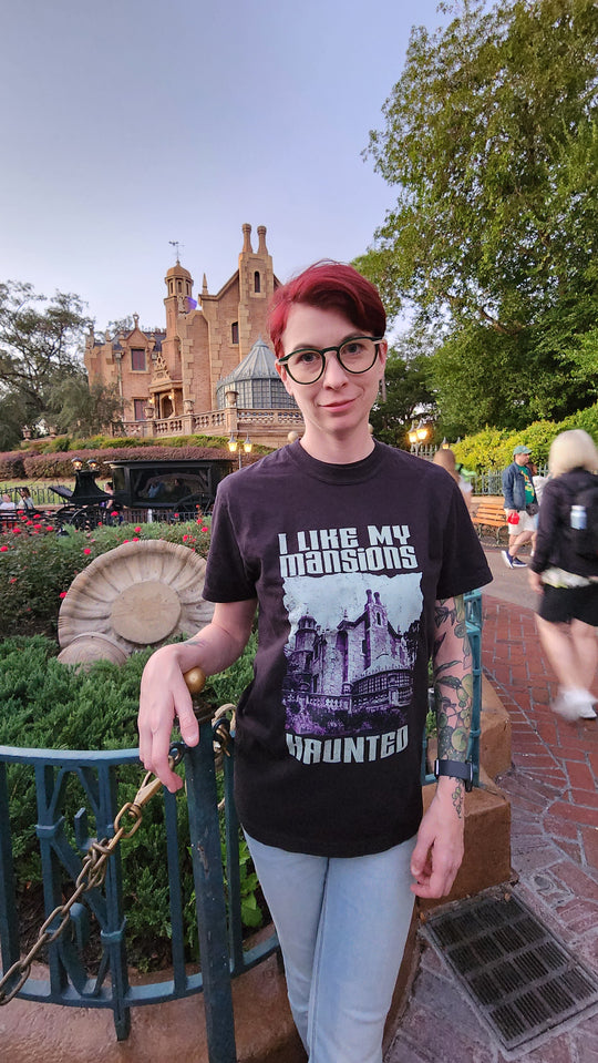 Kylie wearing the I Like My Mansions Haunted Tee in Black with the Haunted Mansion in Liberty Square behind her