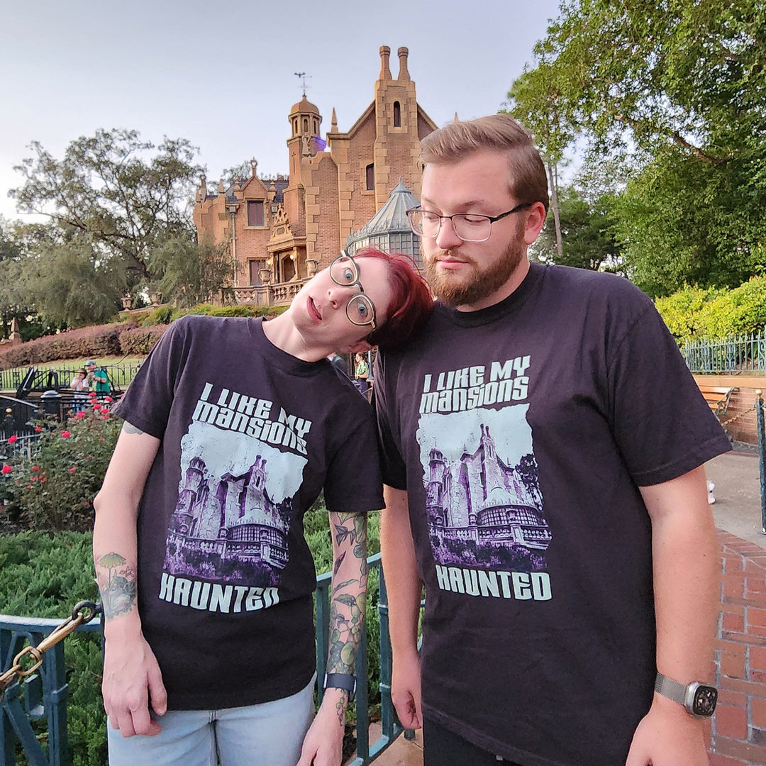 Kylie and a bearded man both wearing the I Like My Mansions Haunted Tee in Black in front of the Haunted Mansion