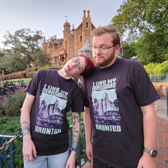 Kylie and a bearded man both wearing the I Like My Mansions Haunted Tee in Black in front of the Haunted Mansion