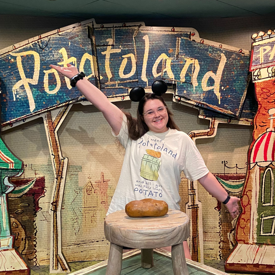 Potatoland Tee in Ivory worn by a woman in Mickey ears, arm raised, beside a potato on a stool at the Potatoland sign