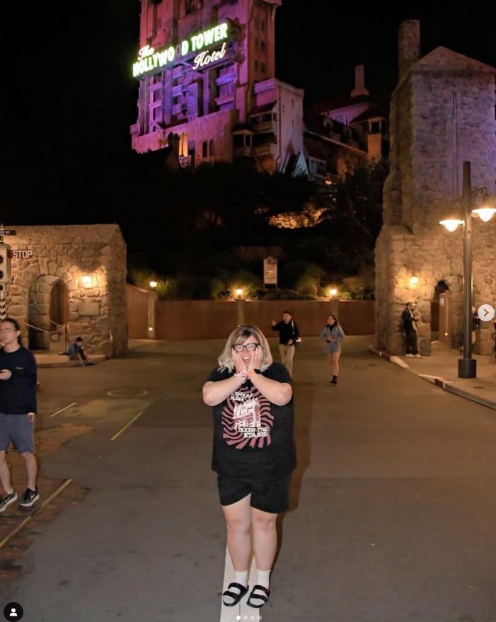 Woman wearing the Should Have Taken The Stairs Tee in Black at night on Sunset Boulevard below the lit Tower of Terror