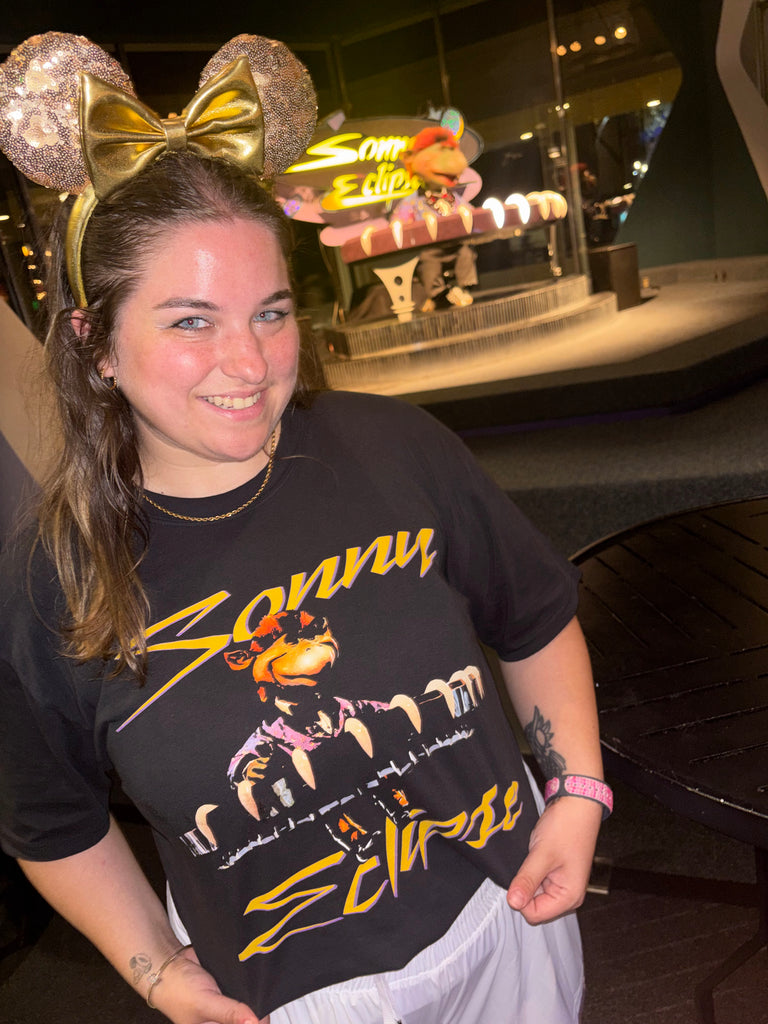 Sonny Eclipse Tee worn by a woman in gold Minnie ears in front of the Sonny Eclipse stage at Cosmic Ray's