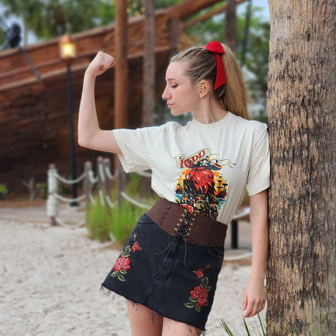 Redd Tee in Ivory worn by a woman with a red hair bow and denim skirt, flexing beside a palm tree
